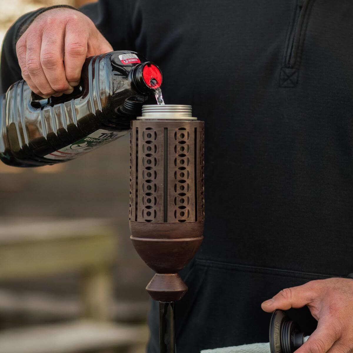 Person pouring TIKI Brand torch fuel into a decorative outdoor torch for backyard or patio use.
