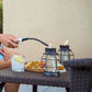 Person lighting TIKI® lanterns on patio table with snacks and a drink, creating a cozy outdoor atmosphere.