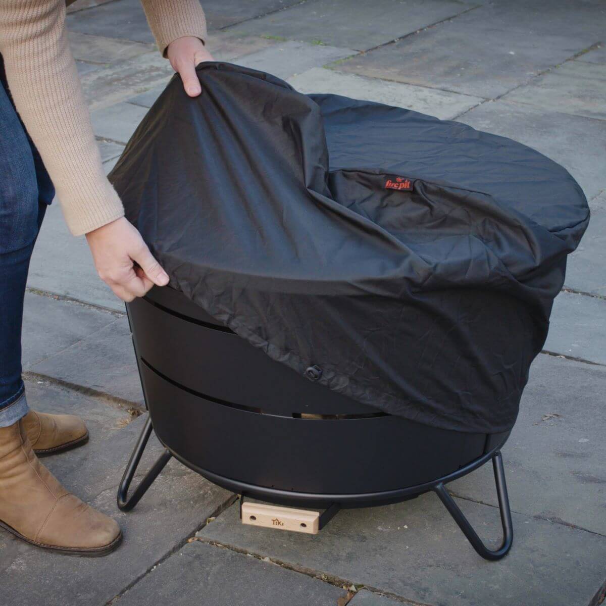 Person covering a TIKI® fire pit with a black weather-resistant cover on a stone patio.