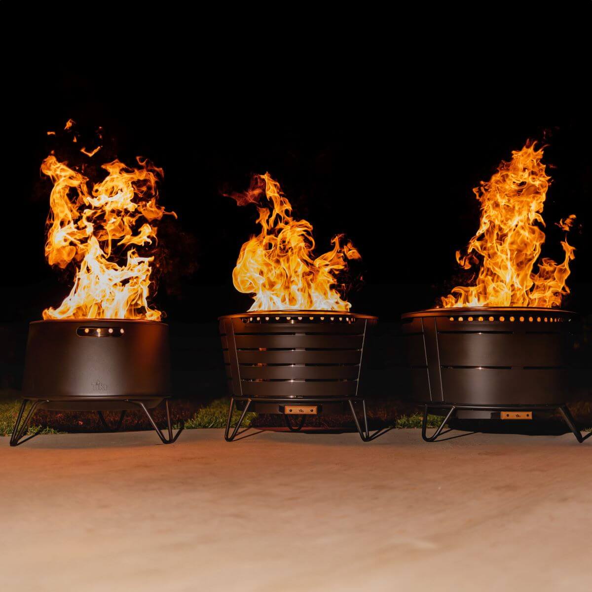 Three black metal fire pits with tall flames, round shape, vent holes, and sturdy legs, shown outdoors at night.
