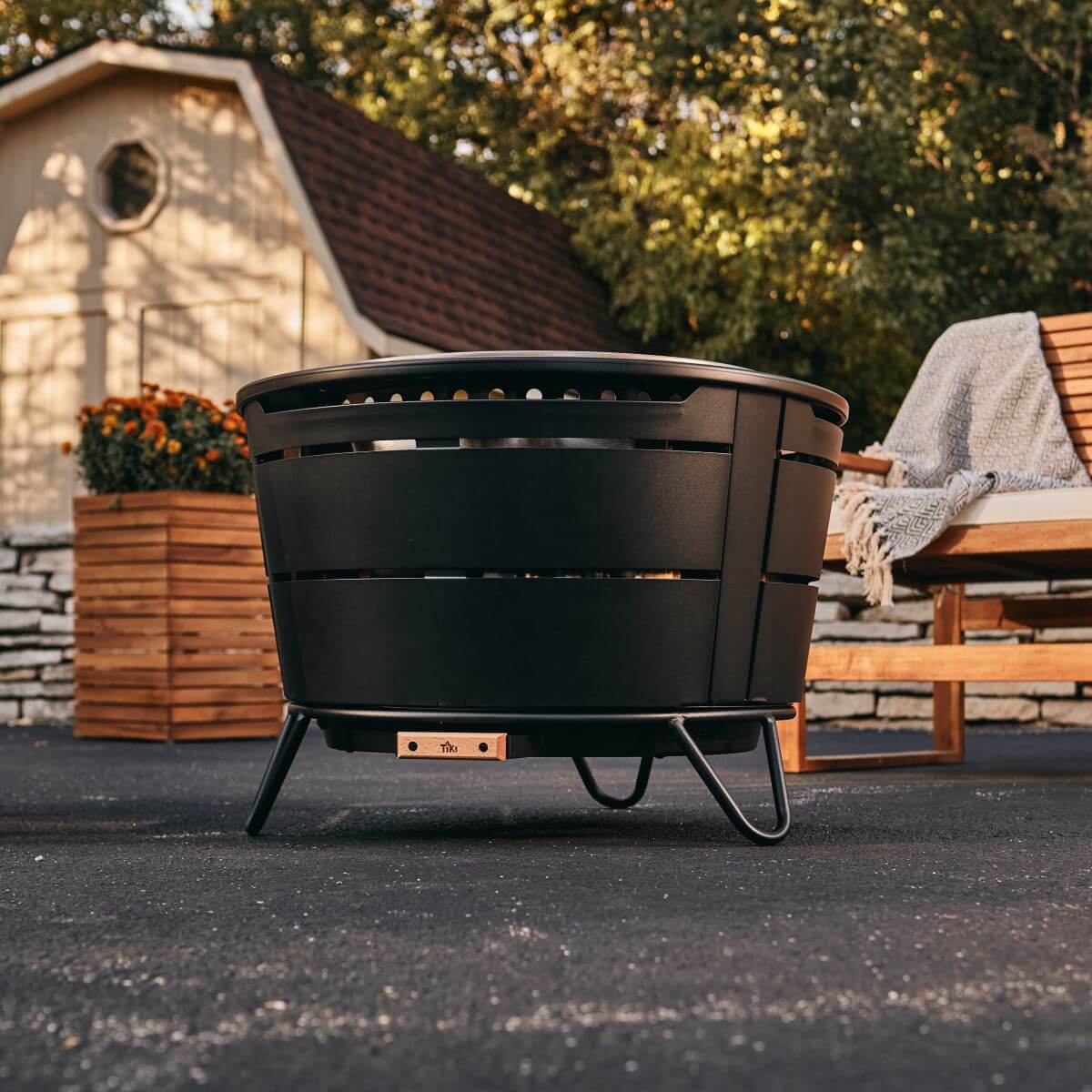 Black TIKI fire pit on patio, near wooden bench and planter, with trees and shed in the background.