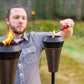 Person lighting a TIKI Brand torch outdoors with a lighter; another torch is already burning in the foreground.