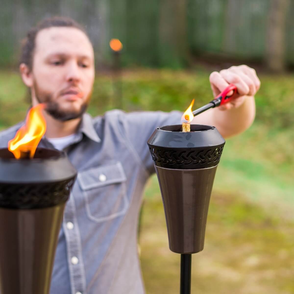 Person lighting a TIKI Brand torch outdoors with a lighter; another torch is already burning in the foreground.