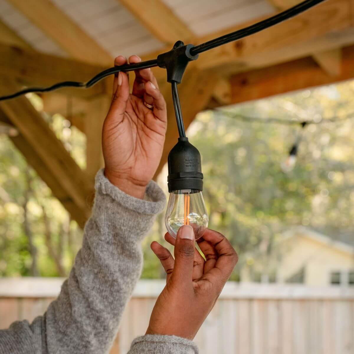 Person installing an outdoor string light bulb under a wooden patio, enhancing backyard ambiance.