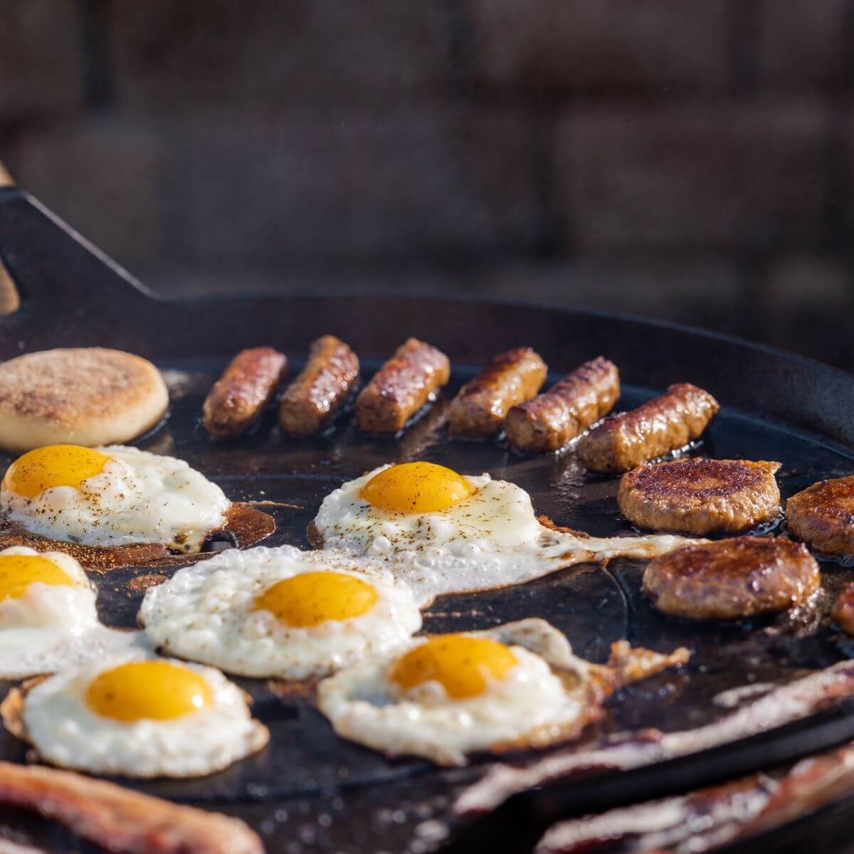 Grilled eggs, sausage links, patties, and an English muffin cooking on a flat-top outdoor griddle.
