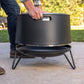 Person lifting a TIKI fire pit by its handle outdoors; fire pit sits on a sturdy metal stand.