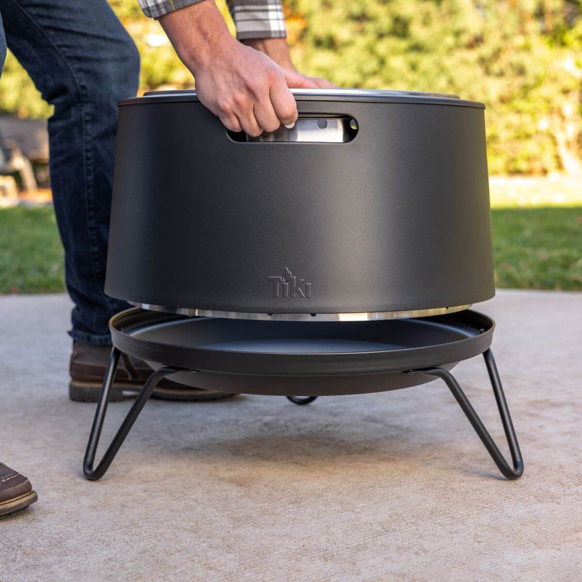 Person lifting a TIKI fire pit by its handle outdoors; fire pit sits on a sturdy metal stand.