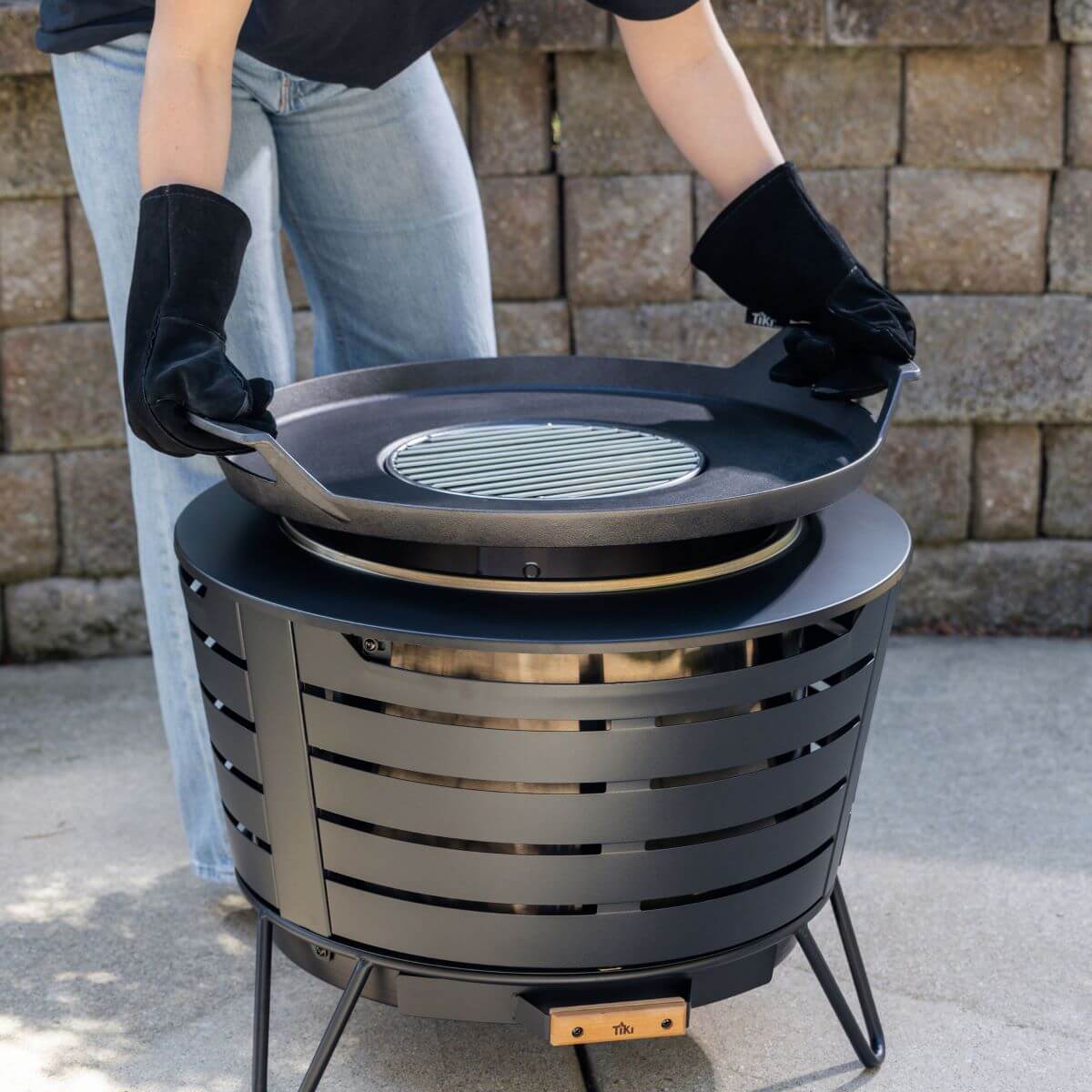 Person wearing gloves lifts grill top from a TIKI Brand fire pit on a patio, with a stone wall in the background.