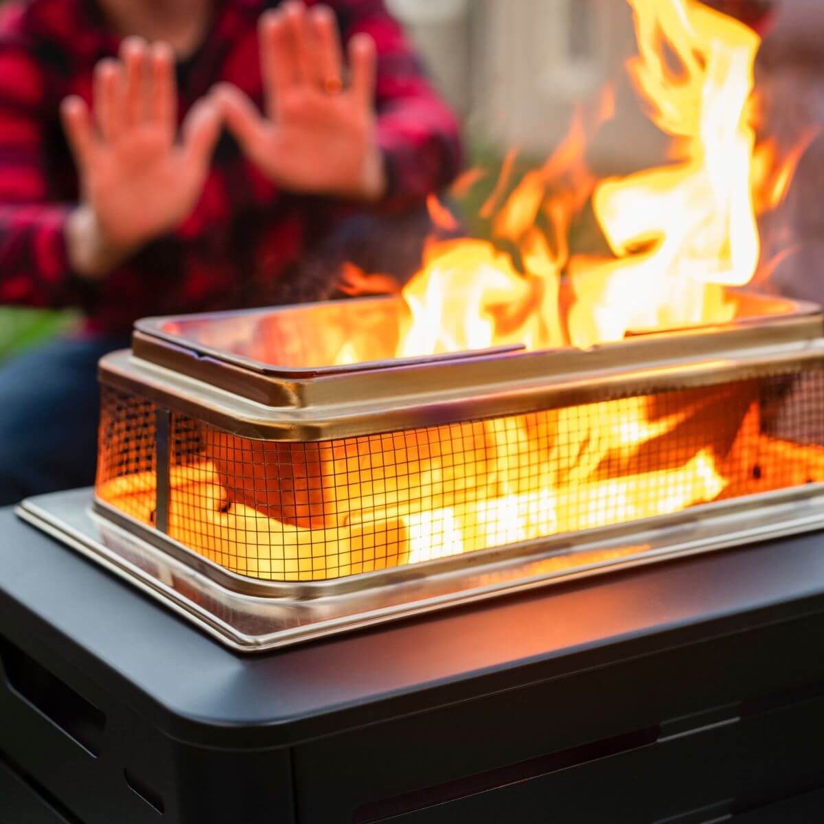 Person warming hands by a rectangular TIKI® fire pit with bright flames, outdoors. Fire pit sits on a dark table.