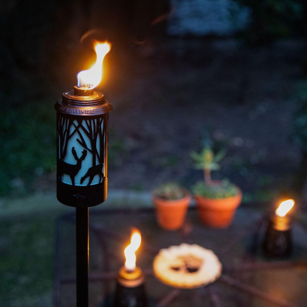 Lit TIKI Brand torch with deer and tree design, glowing in a backyard setting with potted plants and other torches.