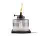 Clear glass TIKI® tabletop torch with visible wick, black metal top, and flame, set on a dark square base.