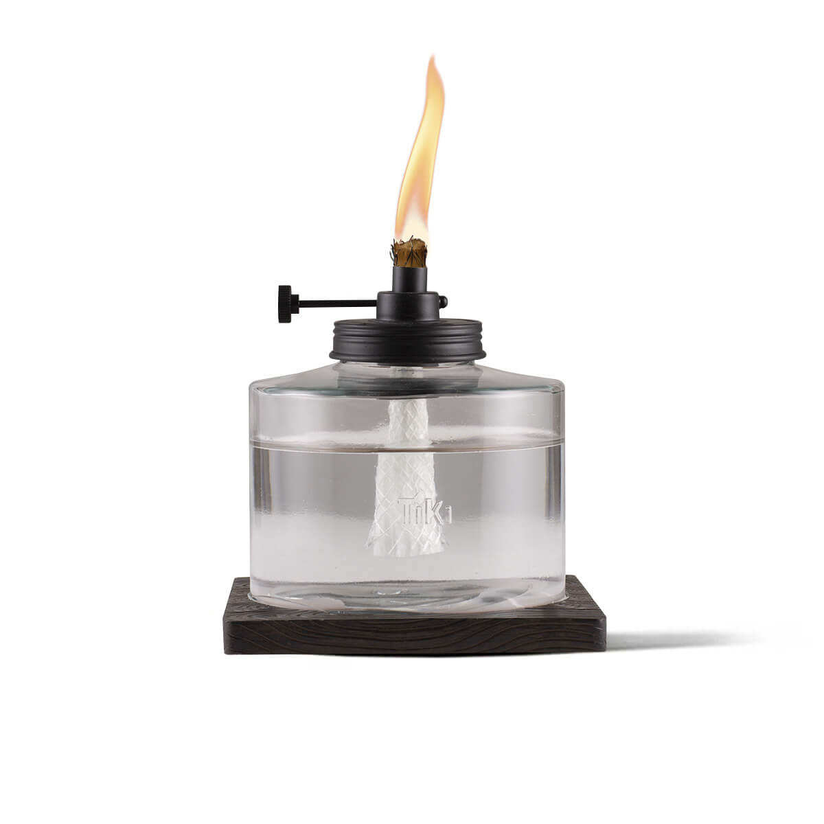 Clear glass TIKI® tabletop torch with visible wick, black metal top, and flame, set on a dark square base.