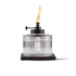 Clear glass TIKI® tabletop torch with visible wick, black metal top, and flame, set on a dark square base.
