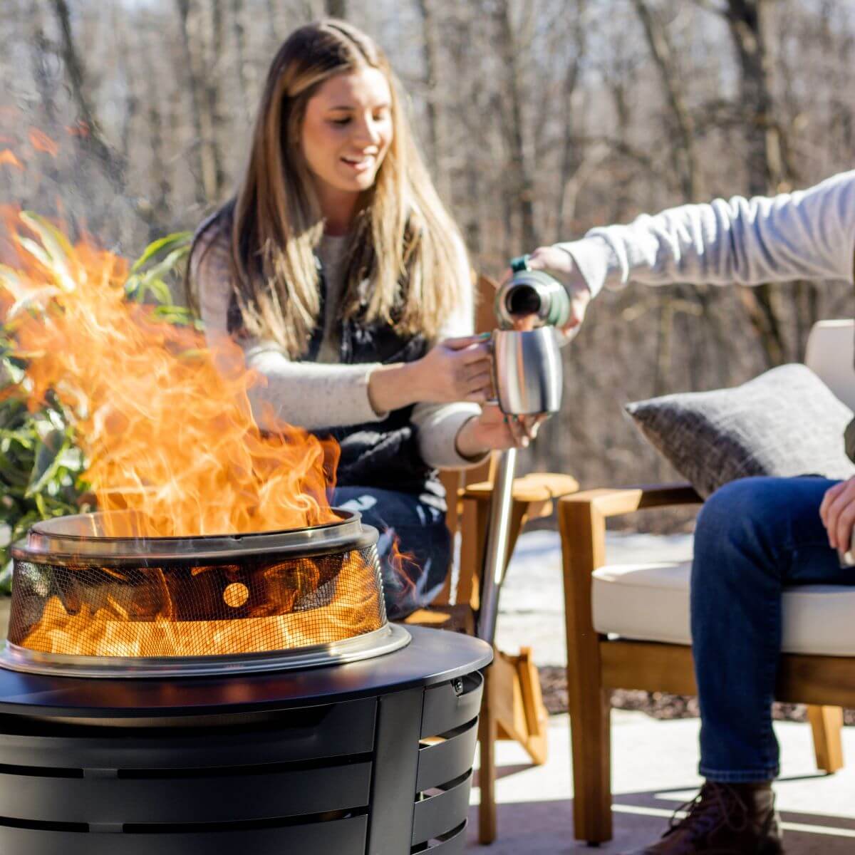 Two people enjoy a warm fire in a TIKI® fire pit on a patio, pouring drinks and relaxing outdoors.