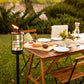 Lit TIKI® lantern beside an outdoor dining table set for a meal, surrounded by greenery in a backyard setting.