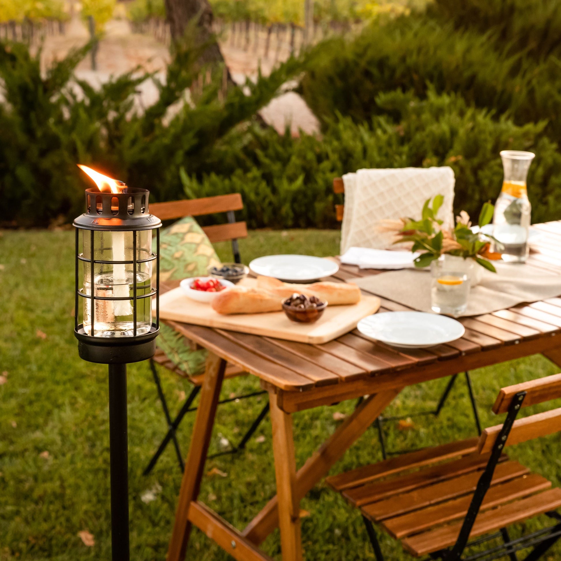 Lit TIKI® lantern beside an outdoor dining table set for a meal, surrounded by greenery in a backyard setting.