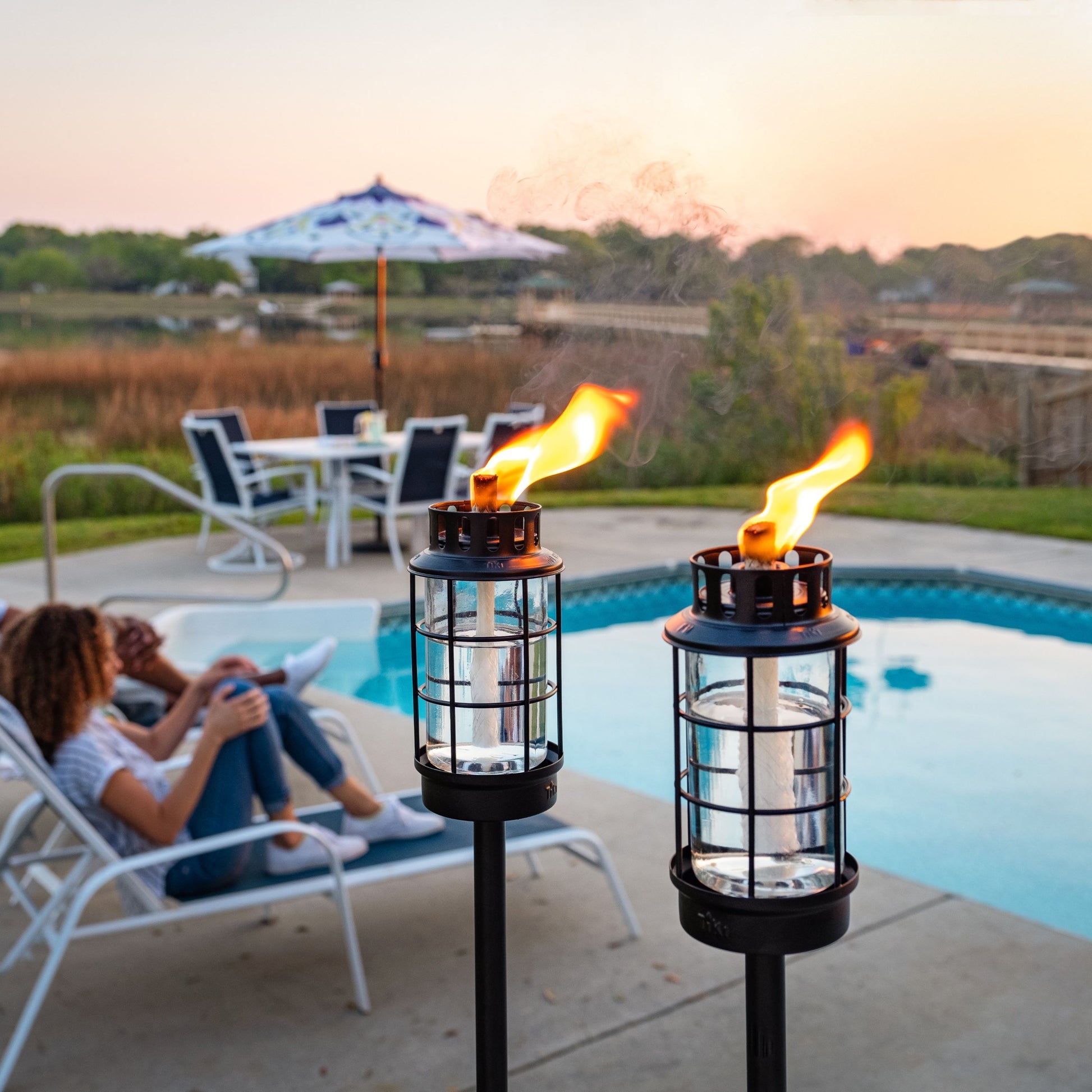 Two TIKI® Brand torches lit by a poolside, creating a cozy outdoor atmosphere with patio seating and scenic views.