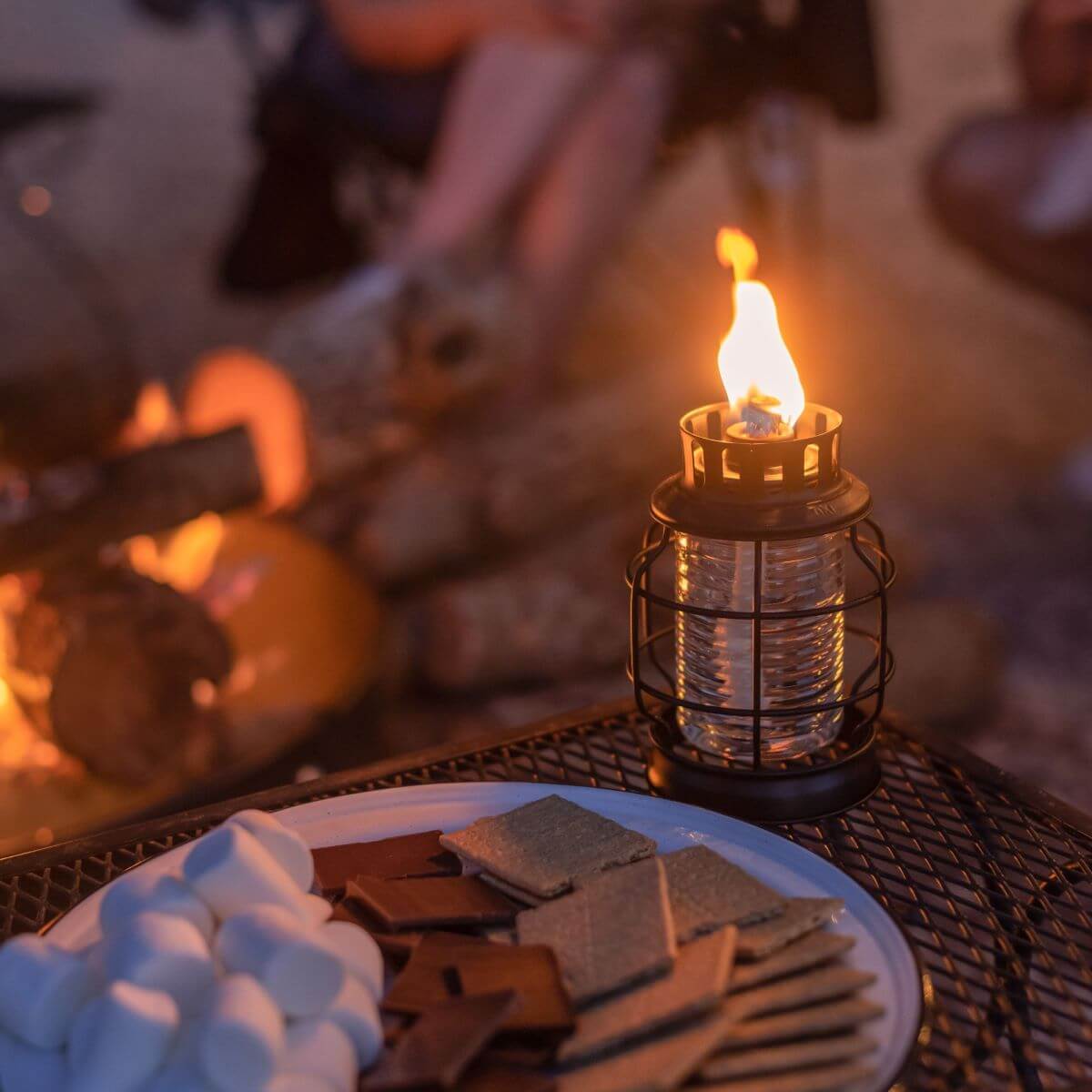 Plate of s'mores ingredients beside a lit TIKI® lantern on an outdoor table, with a campfire in the background.