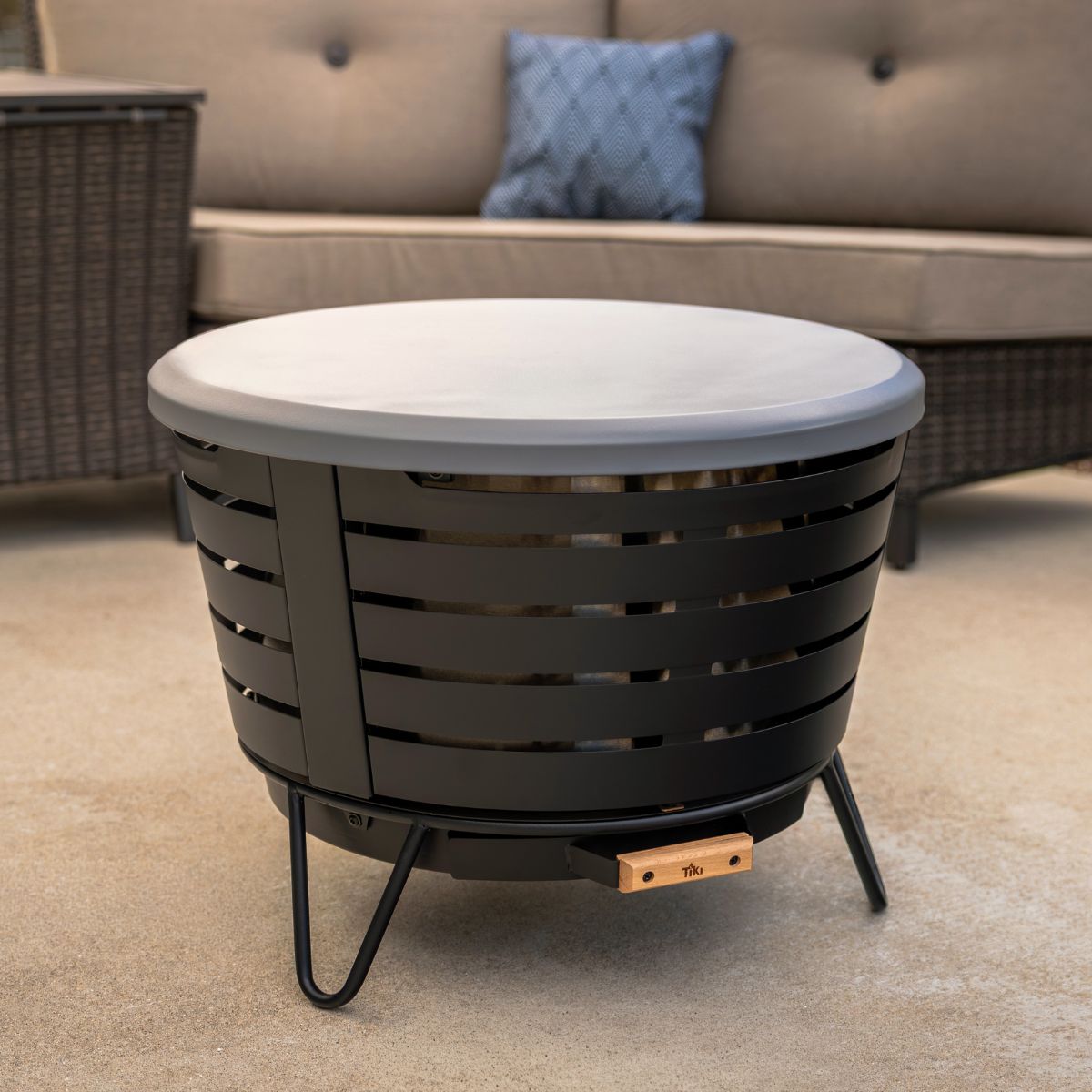 Round black metal fire pit with a flat lid, slatted sides, and sturdy legs, placed on a patio.