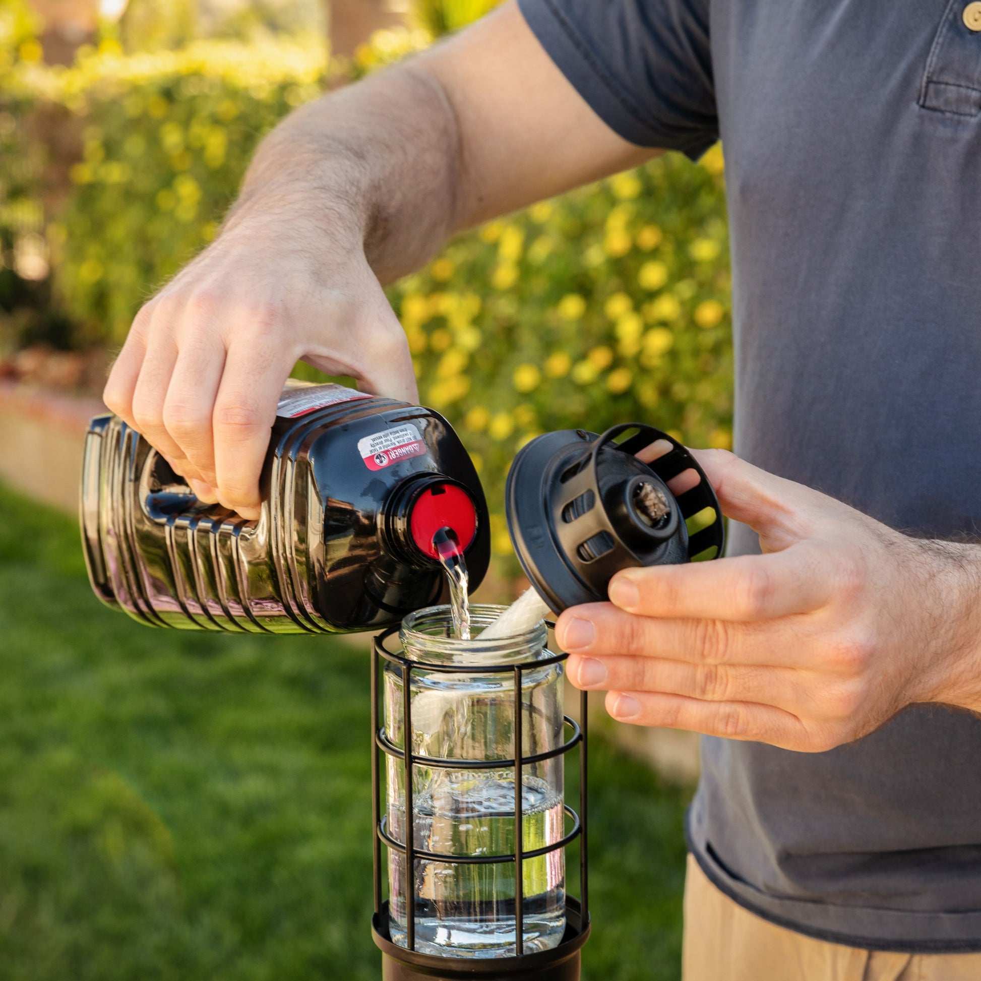 Person pours TIKI torch fuel into a torch canister outdoors, preparing for safe backyard lighting.