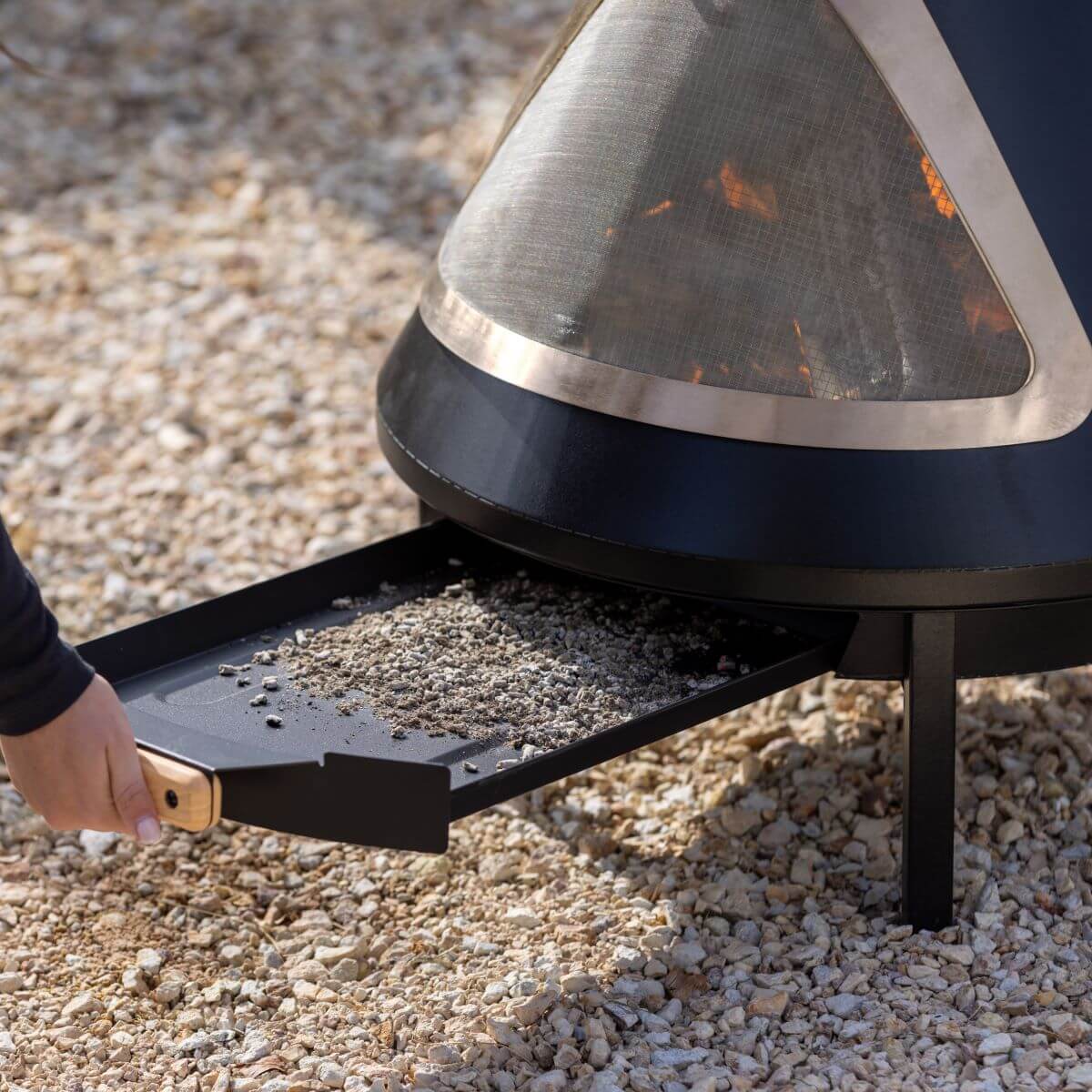 A hand pulls out an ash tray from a TIKI Brand fire pit for easy cleaning on a gravel surface.