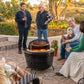 Group enjoying drinks and conversation around a TIKI Brand fire pit on a patio, with flowers and a dog nearby.