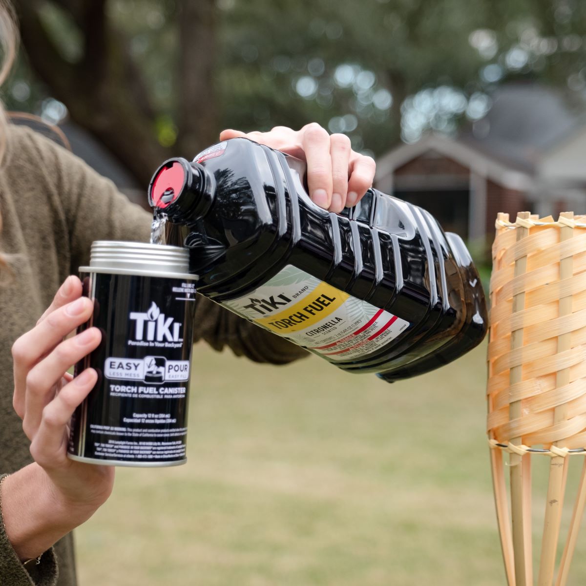 Person pours TIKI Brand torch fuel into an easy-pour canister outdoors, next to a bamboo torch.