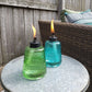 Two TIKI® glass table torches, green and blue, lit on a patio table beside a wicker chair with cushions.