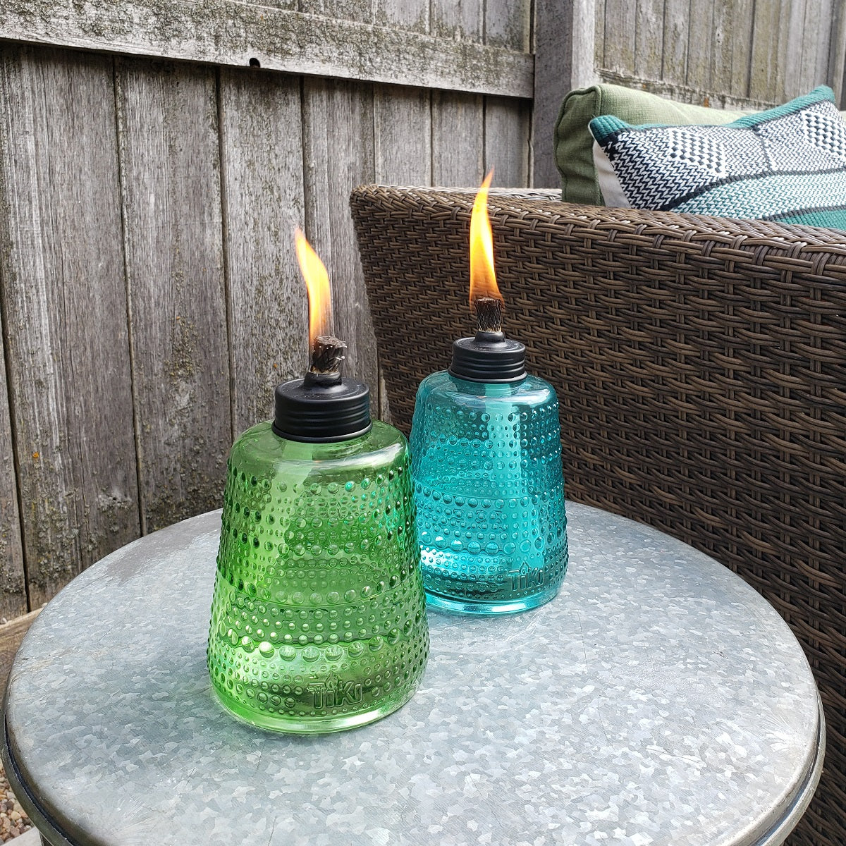 Two TIKI® glass table torches, green and blue, lit on a patio table beside a wicker chair with cushions.