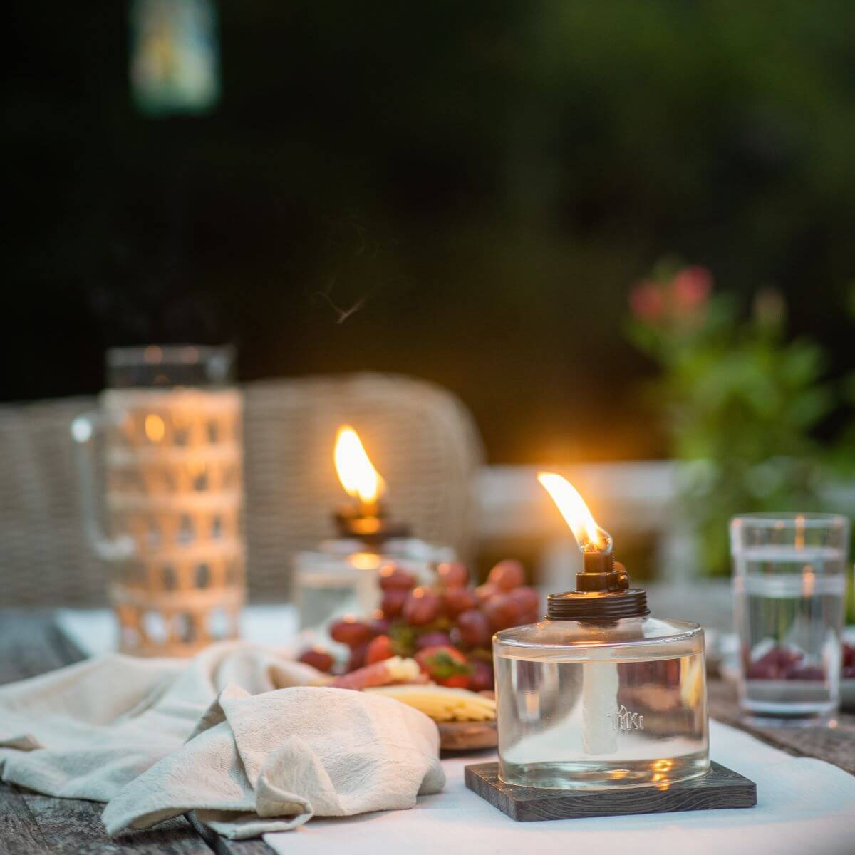 Two TIKI® tabletop torches with bright flames on an outdoor dining table set with fruit, water, and a napkin.