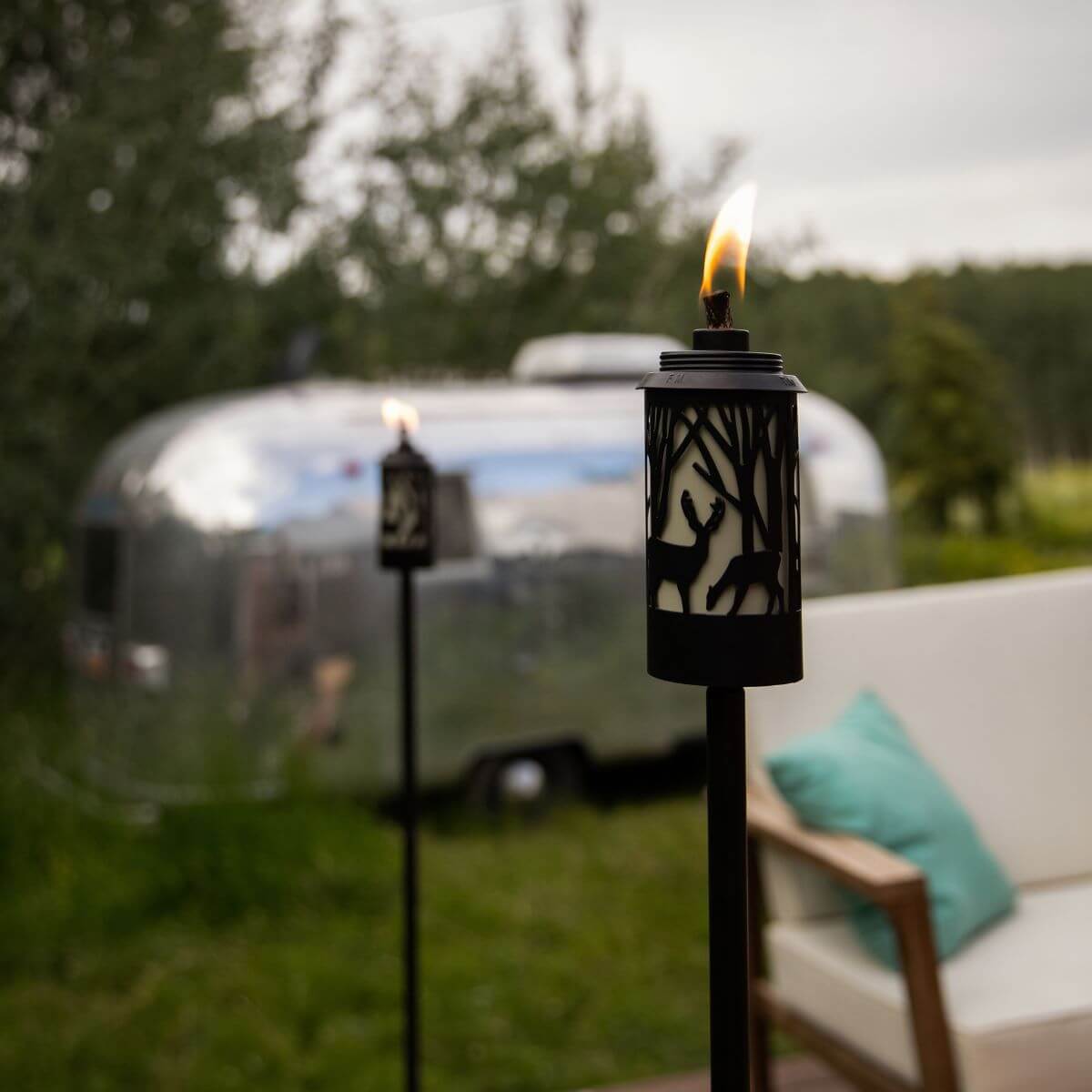 Lit TIKI Brand torches with deer design on a patio, near a camper and outdoor seating. Accessible outdoor ambiance.