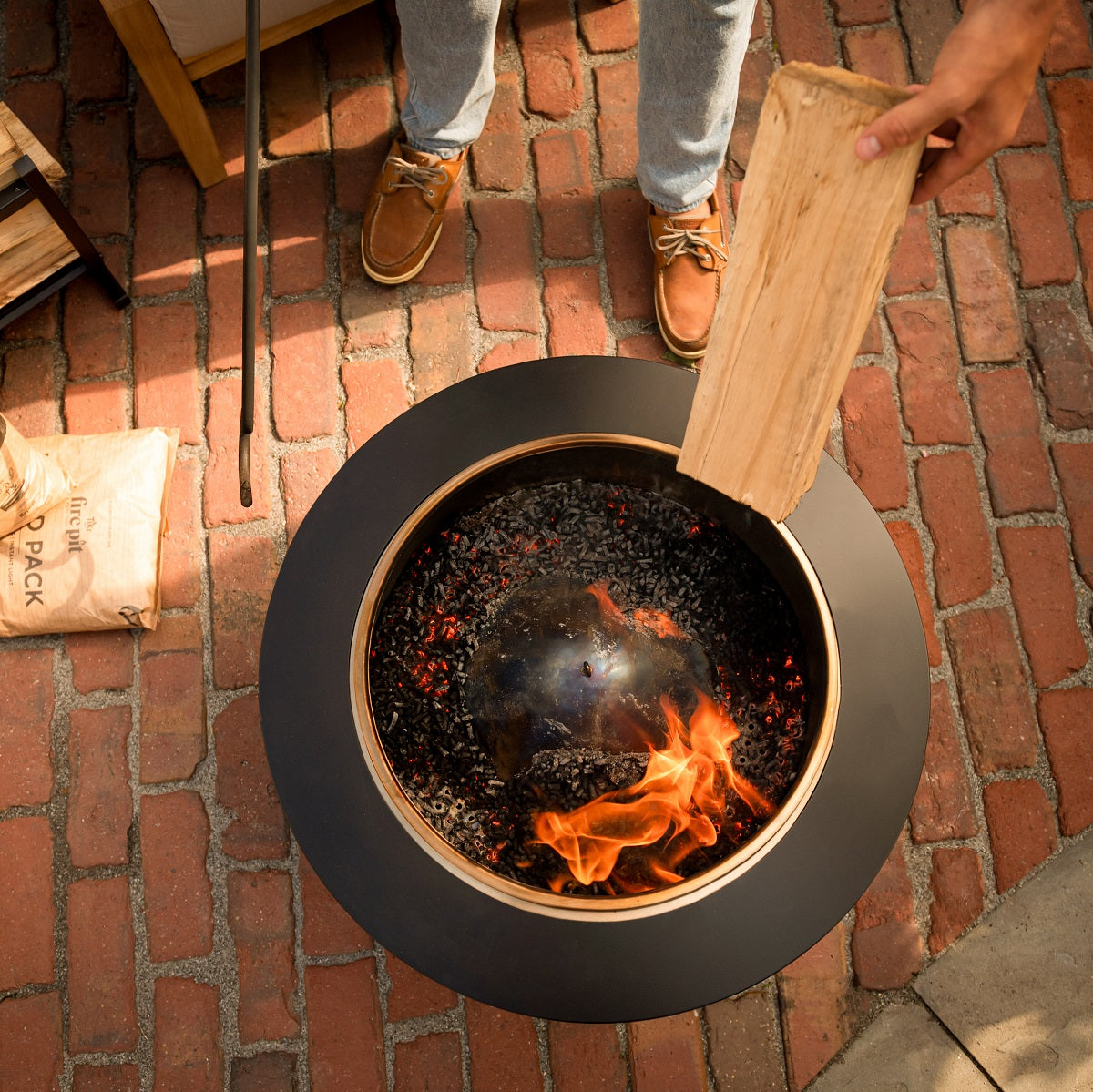 Person adding a wood log to a TIKI® fire pit on a brick patio, with flames and embers visible inside the fire pit.