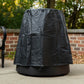 Black weather-resistant cover protects a TIKI® fire pit on a patio, with trees and a brick wall in the background.