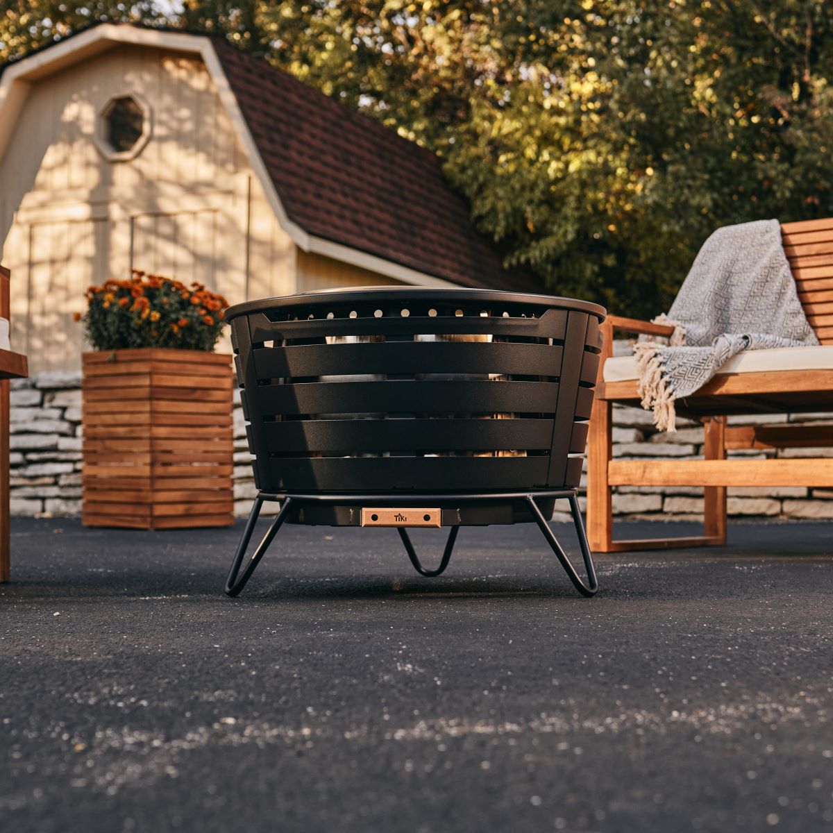 Black metal outdoor fire pit with slatted sides and sturdy legs, placed on patio near wooden seating and planter.