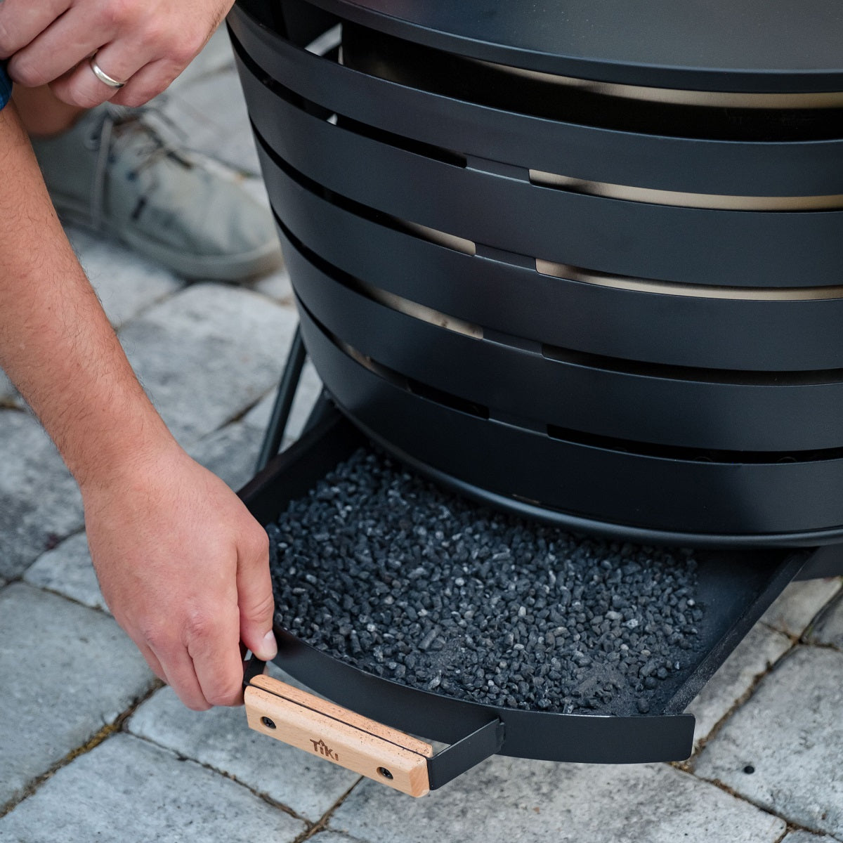 Black metal fire pit with a removable ash tray, filled with charcoal, on a stone patio. Wooden handle for easy grip.