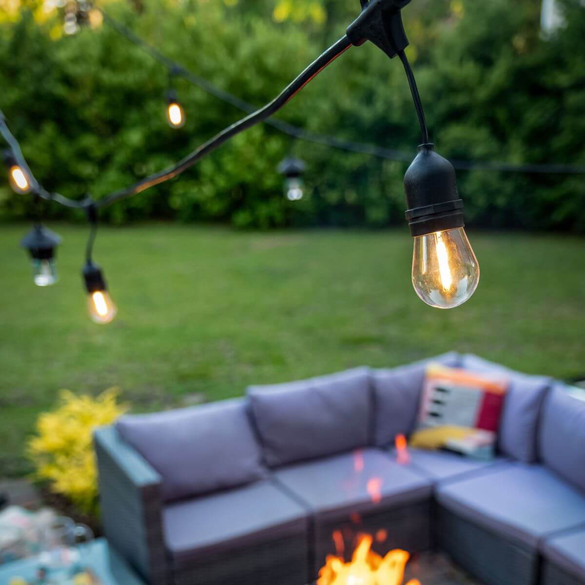 String lights hang above an outdoor patio with a gray sectional sofa and fire pit, surrounded by green lawn and bushes.