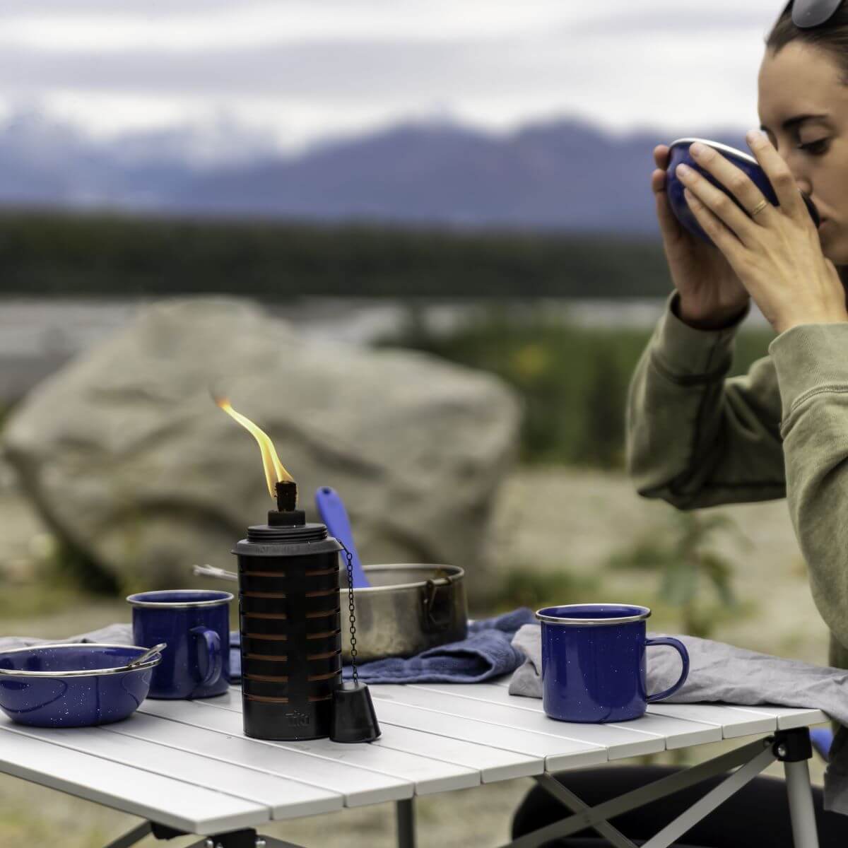 A tabletop TIKI® torch burns beside blue camping mugs and bowls on an outdoor table with a scenic mountain backdrop.