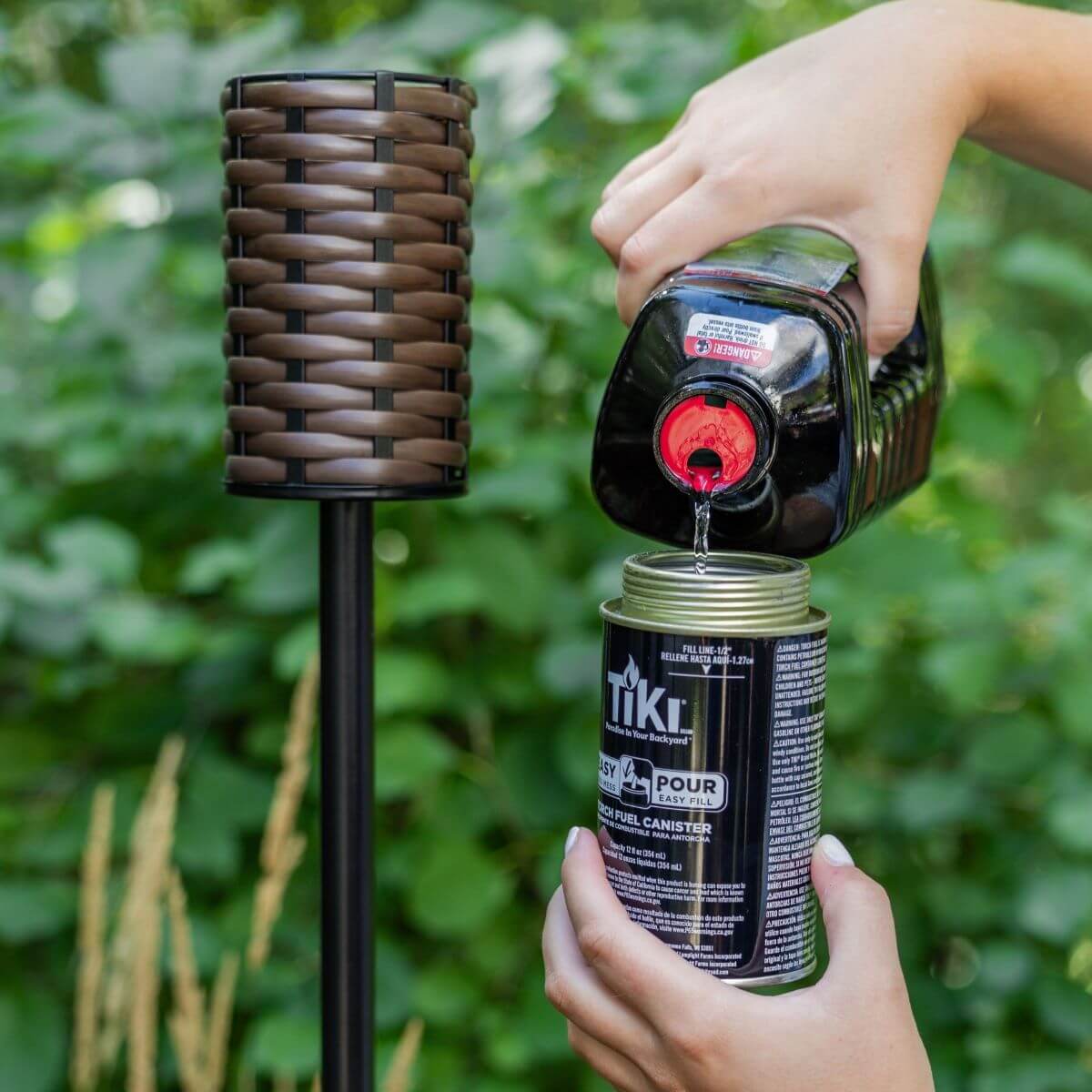 Person pouring TIKI torch fuel into a canister outdoors, preparing a woven brown TIKI torch for use.