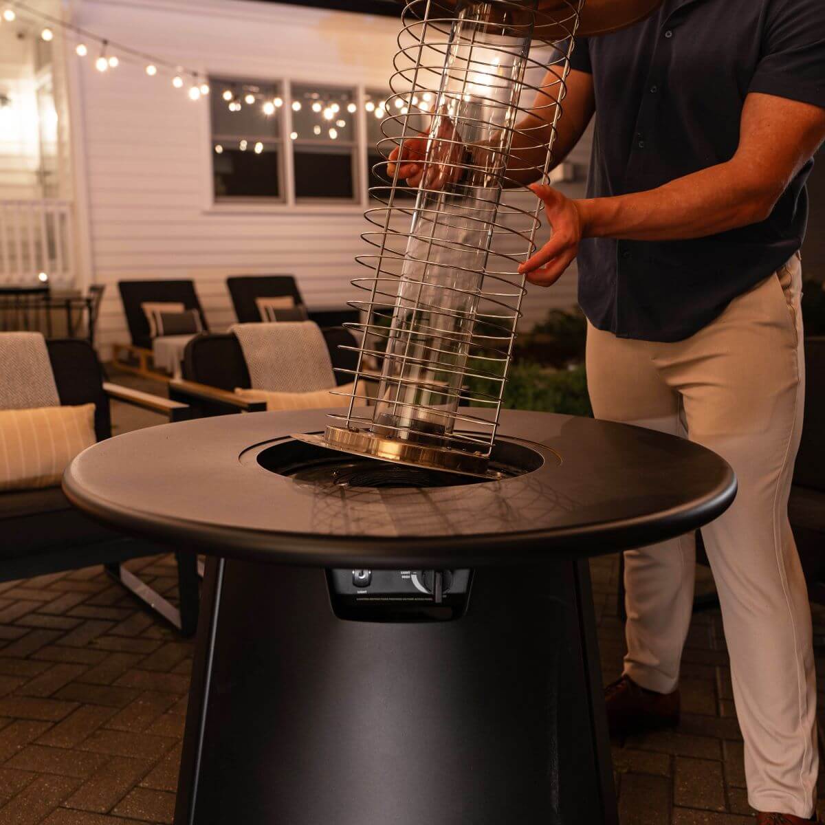 A person installs a metal fire feature into a TIKI® patio fire pit table on an outdoor patio at night.
