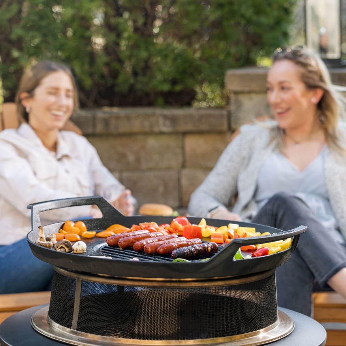 Outdoor TIKI fire pit grill with sausages and vegetables cooking, surrounded by people relaxing in a backyard setting.