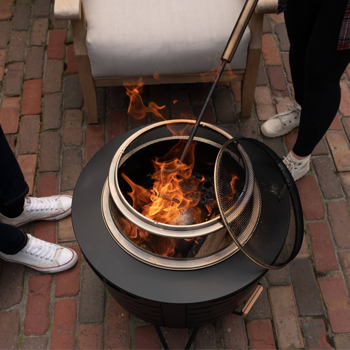 Two people tend a fire in a TIKI® fire pit on a brick patio, using a poker and mesh screen for safety.