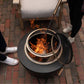 Black outdoor fire pit with mesh cover and poker, flames visible.