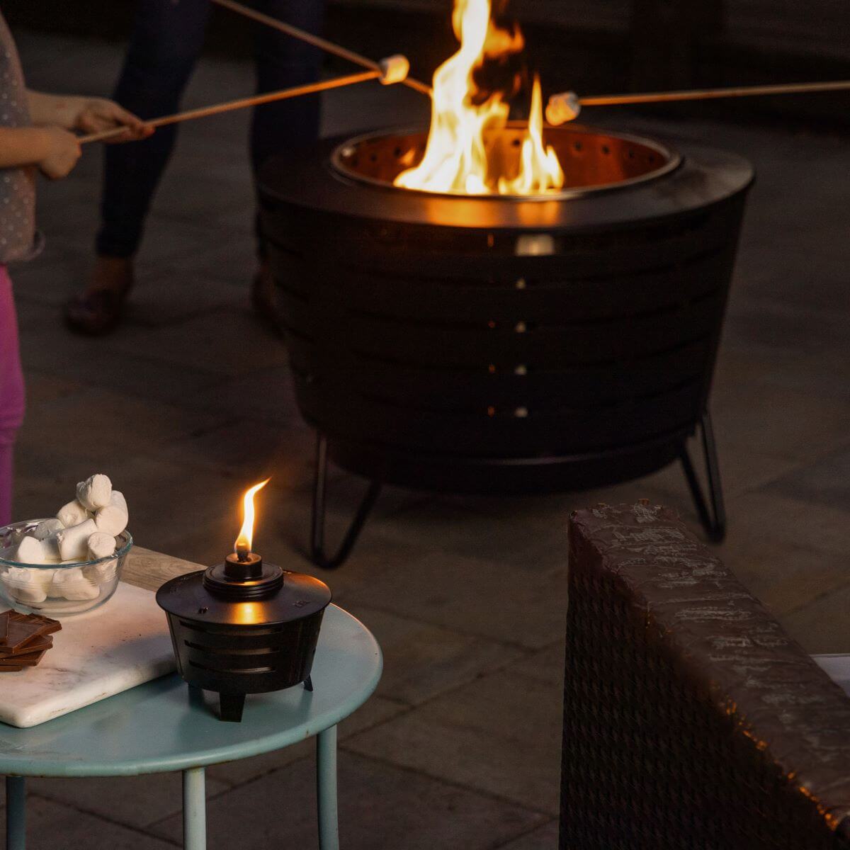 Family roasting marshmallows over a TIKI® fire pit, with a tabletop torch and s'mores ingredients nearby.