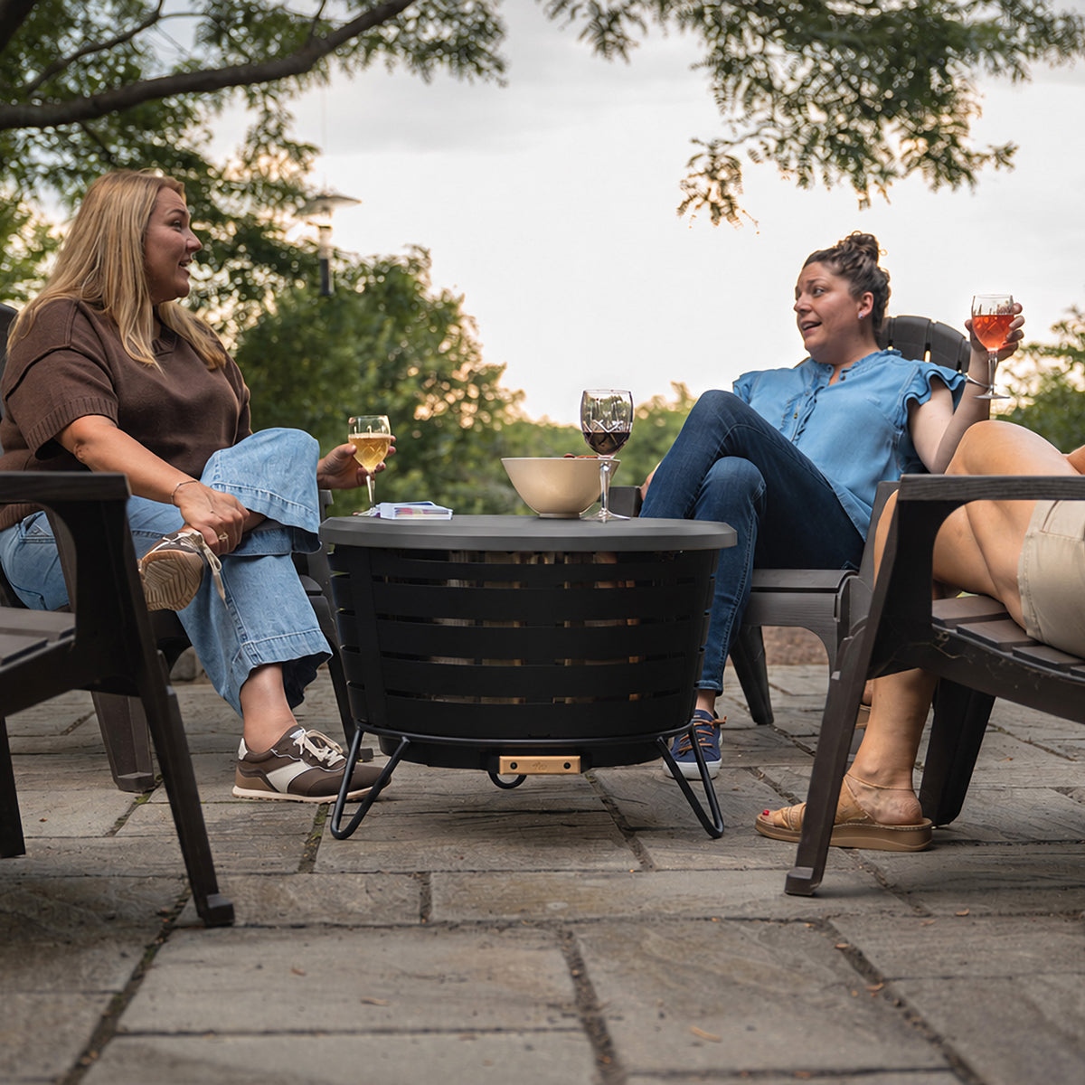 Round black outdoor fire pit table with slatted sides, set on a patio with drinks and snacks on top.