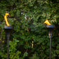 Pair of black outdoor torches with patterned tops, each featuring a bright flame against a leafy green background.