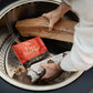 Person places logs and a TIKI Fire Starter pack inside a fire pit, preparing to start a wood fire easily and safely.