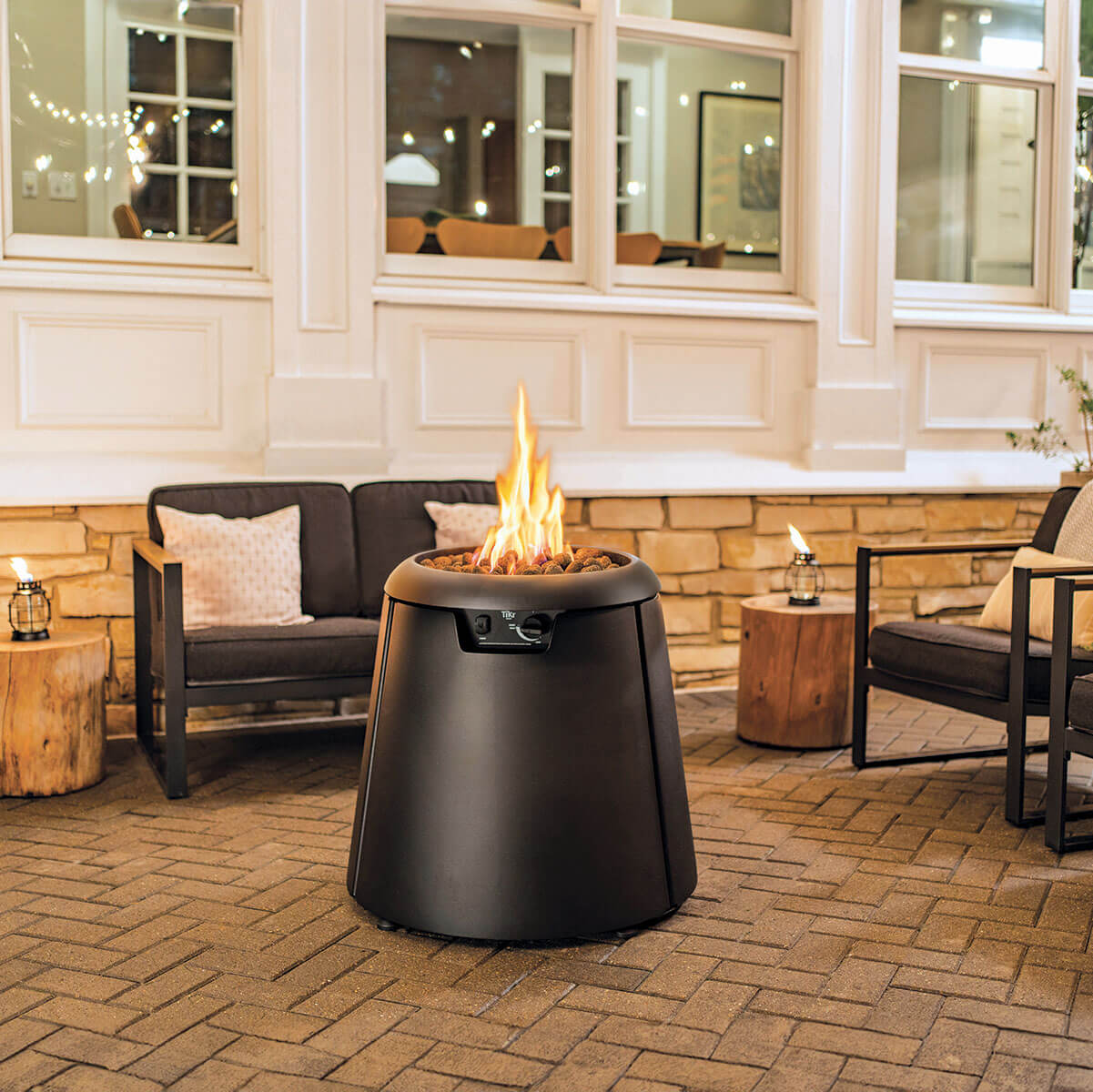 Modern black TIKI® fire pit with visible flame on a brick patio, surrounded by outdoor seating and soft lighting.