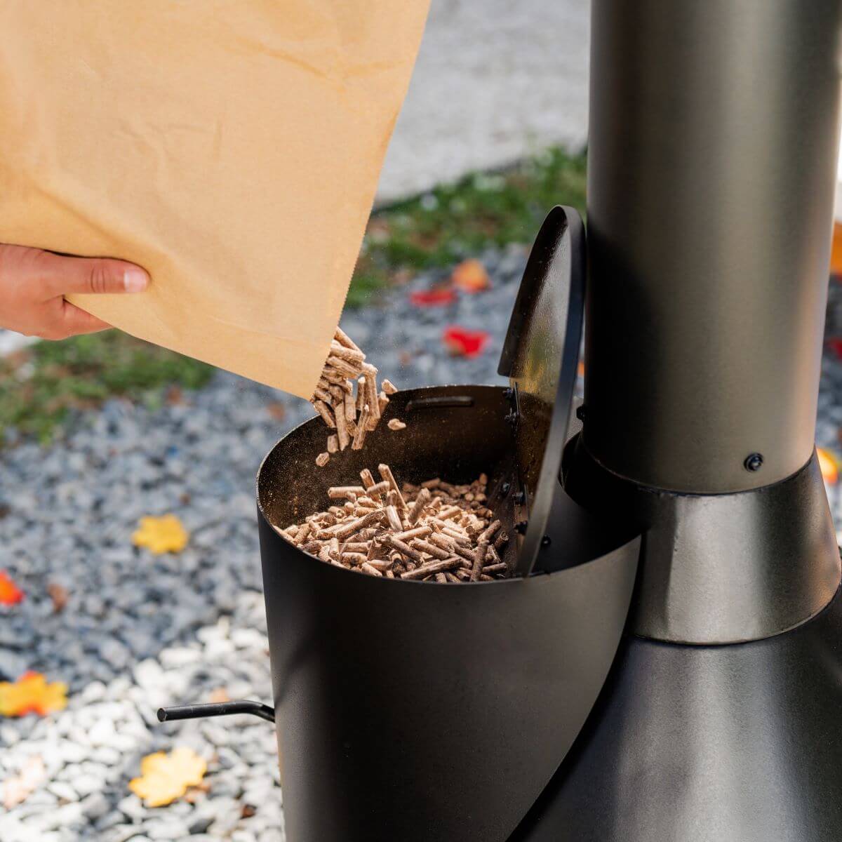 Hand pouring wood pellets into a TIKI Brand fire pit hopper outdoors, preparing for a smokeless fire.