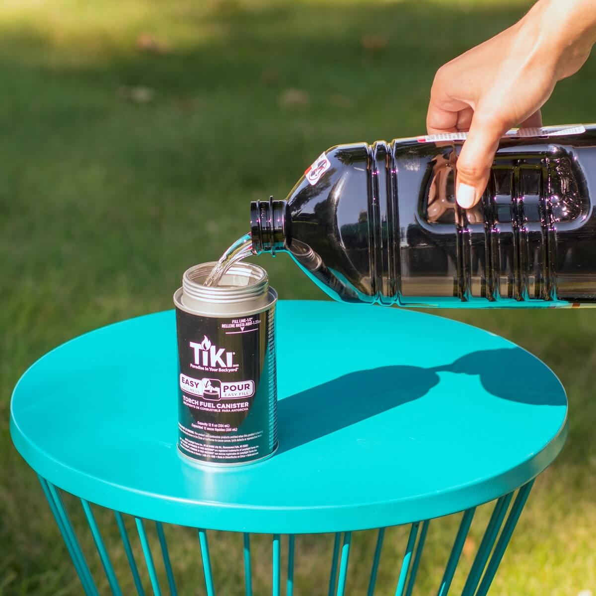 A hand pours TIKI torch fuel into an Easy Pour canister on a teal outdoor table, with grass in the background.