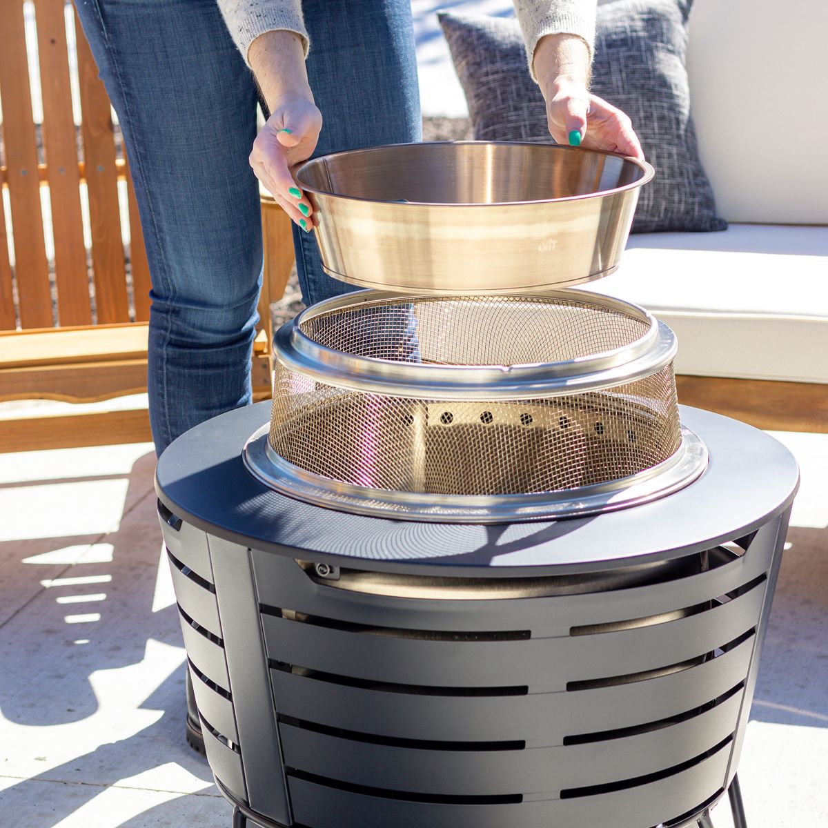 Person placing a metal bowl onto a TIKI fire pit outdoors, with patio furniture in the background.